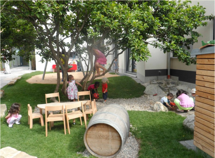 8 Kinder spielen auf und um einen Baum herum. Auf der Wiese stehen ein Holztisch und Stühle für Kinder. Daneben liegt ein Holzfass im Gras.