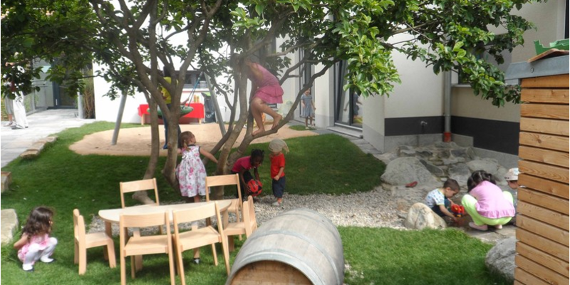 8 Kinder spielen auf und um einen Baum herum. Auf der Wiese stehen ein Holztisch und Stühle für Kinder. Daneben liegt ein Holzfass im Gras.