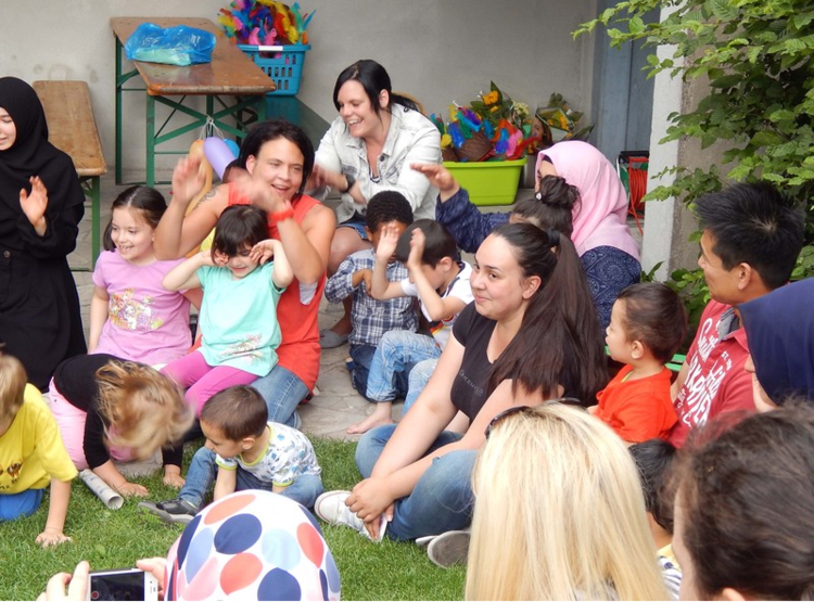 Ca. 20 Kinder und Erwachsene sitzen auf einer Wiese. Viele lachen, einige bewegen ihre Hände. Im Hintergrund Kisten mit bunten Spielsachen. 