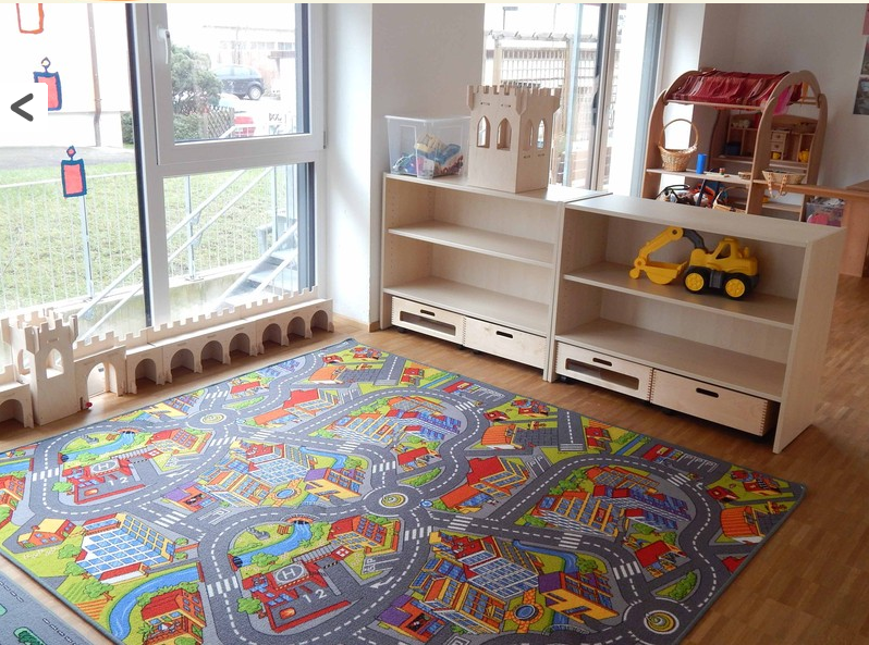 Ein Spielzimmer mit einem Verkehrsteppich. In einem Holzregal lagern ein gelber Bagger, ein Holzturm und eine Plastikbox mit bunten Spielsachen.