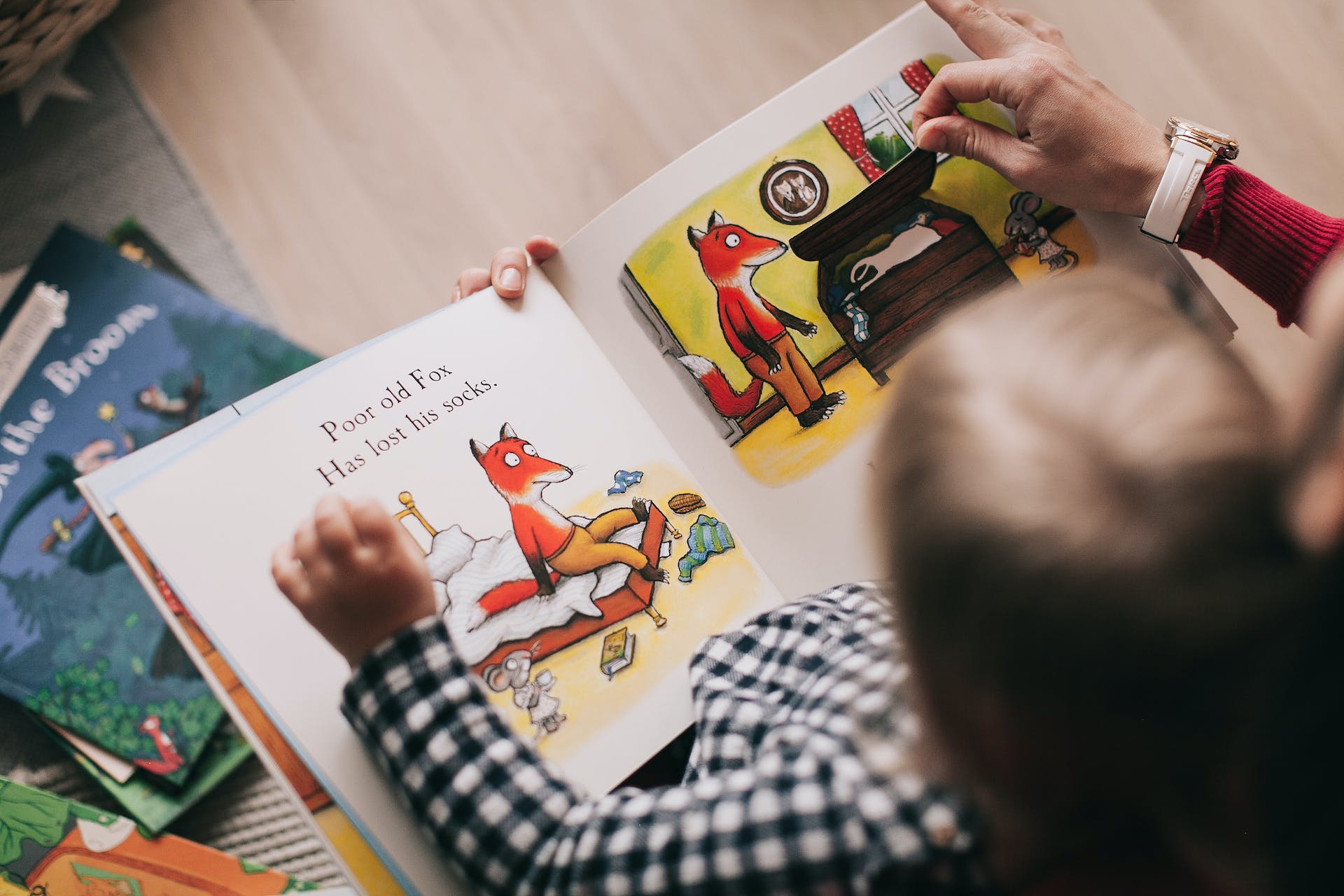 Das Buch "Poor old Fox has lost his socks" liegt auf dem Schoß eines Kindes, das wiederum auf dem Schoß einer Erwachsenen sitzt. Die Hand der Erwachsenen öffnet eine Truhe im Buch.
